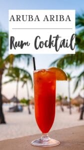 A tall glass of red rum cocktail with an orange slice and straw, set outdoors against a beach backdrop with palm trees, features the "Aruba Ariba" text overlay.