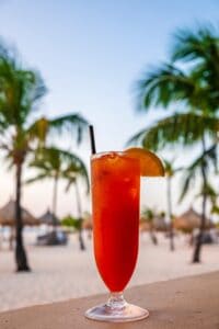 A tall glass of the vibrant Aruba Ariba cocktail with ice and a slice of orange sits on a ledge, framed by palm trees and a sandy beach in the background, all under a clear sky.