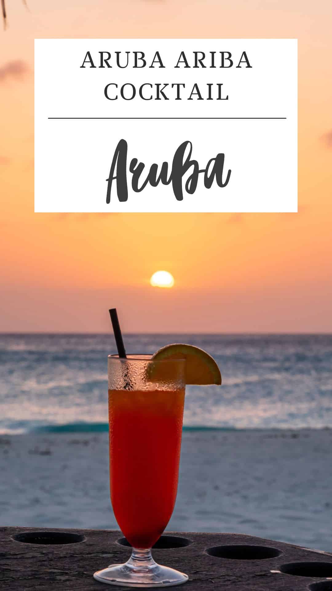 A red Aruba Ariba cocktail with a lime wedge sits on a table by the beach at sunset, with the text "Aruba Ariba Cocktail Aruba" above.