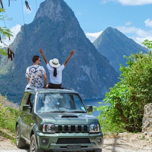 A couple enjoys a scenic mountain view sitting on the roof of a green SUV, with the woman raising her arms joyfully under a clear sky, showcasing how to get around St. Lucia.