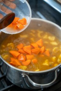 Carrot pieces are carefully added to a simmering pot of soup on the stove, enriched with savory pig tail bouillon.