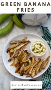 A plate of crispy green banana fries with a side of dipping sauce rests on a white surface alongside whole green bananas and a stylish green striped cloth.