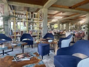 The modern restaurant interior, perfect after a day of shopping in Saint Lucia, features blue chairs and wooden tables. A shelving unit holds bottles, while circular light fixtures illuminate the dining tables set for a delightful experience.