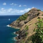 Rocky, grassy headland with patches of green vegetation extends into blue ocean under a partly cloudy sky; a white sailboat is visible in the distance, capturing the scenic beauty of the Pigeon Island hike.