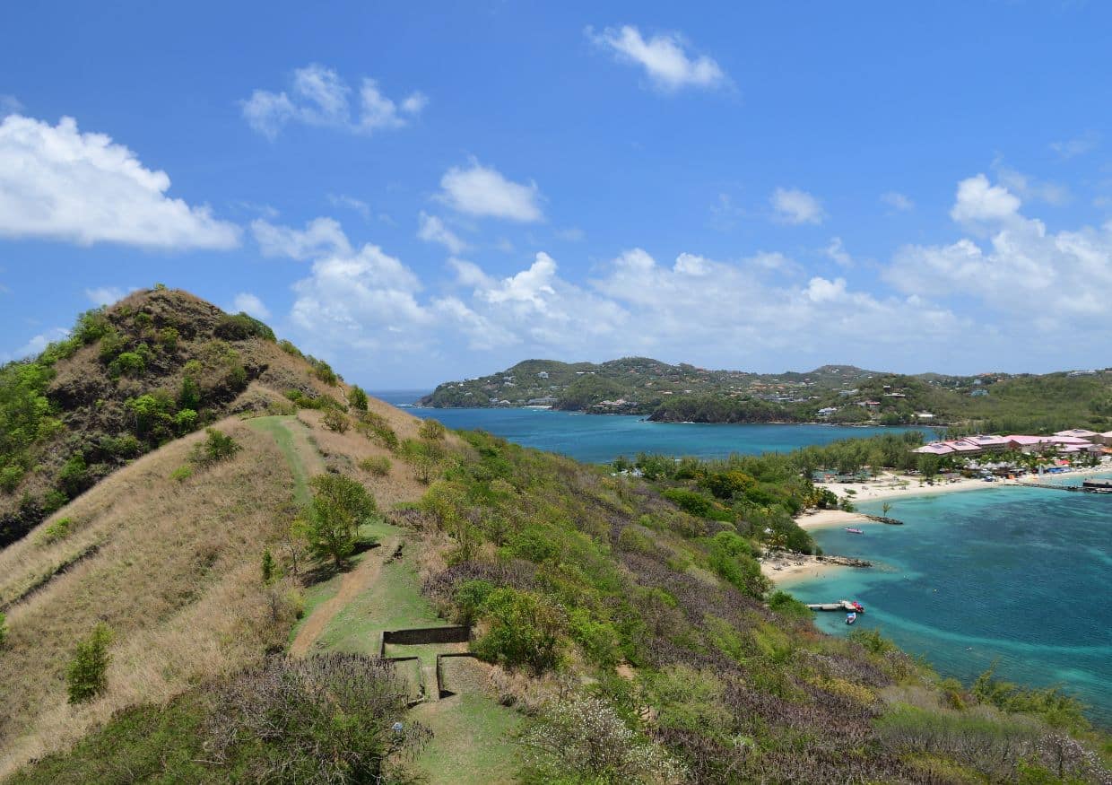 A coastal landscape with a grassy hill on the left, clear blue water, scattered boats, and buildings along the shoreline under a bright sky—perfect scenery for those enjoying the pigeon island hike nearby.