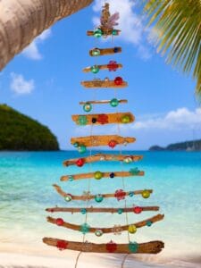A Christmas tree made of driftwood and ornaments hangs by the beach, capturing the essence of Christmas in St Lucia. Turquoise waters and islands shimmer in the background, while palm fronds elegantly frame this tropical holiday scene.