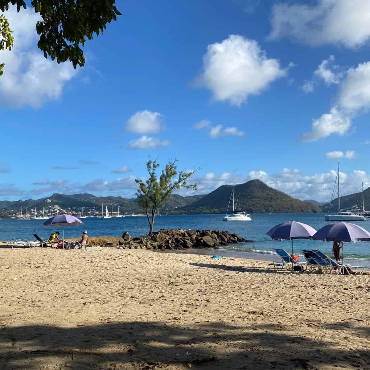 Sandy beach with lounge chairs and umbrellas on Pigeon Island, St. Lucia. People relax near the water as sailboats are anchored in the bay. Hills rise underneath a blue sky adorned with scattered clouds in the background.