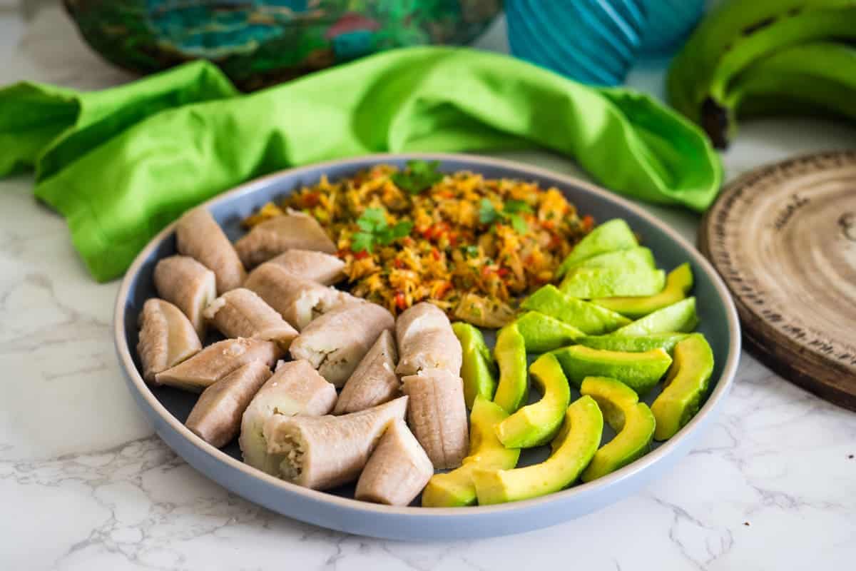 A plate with boiled plantains, sliced avocado, and spicy rice garnished with cilantro is set on a marble surface with green fabric in the background, echoing flavors of Caribbean cuisine where saltfish often pairs perfectly.