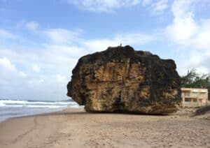 Explore this large rock formation on a sandy beach, where ocean waves crash under partly cloudy skies. Among the top things to do in Barbados, you'll spot a charming building to the right, adding a touch of island allure to this scenic view.