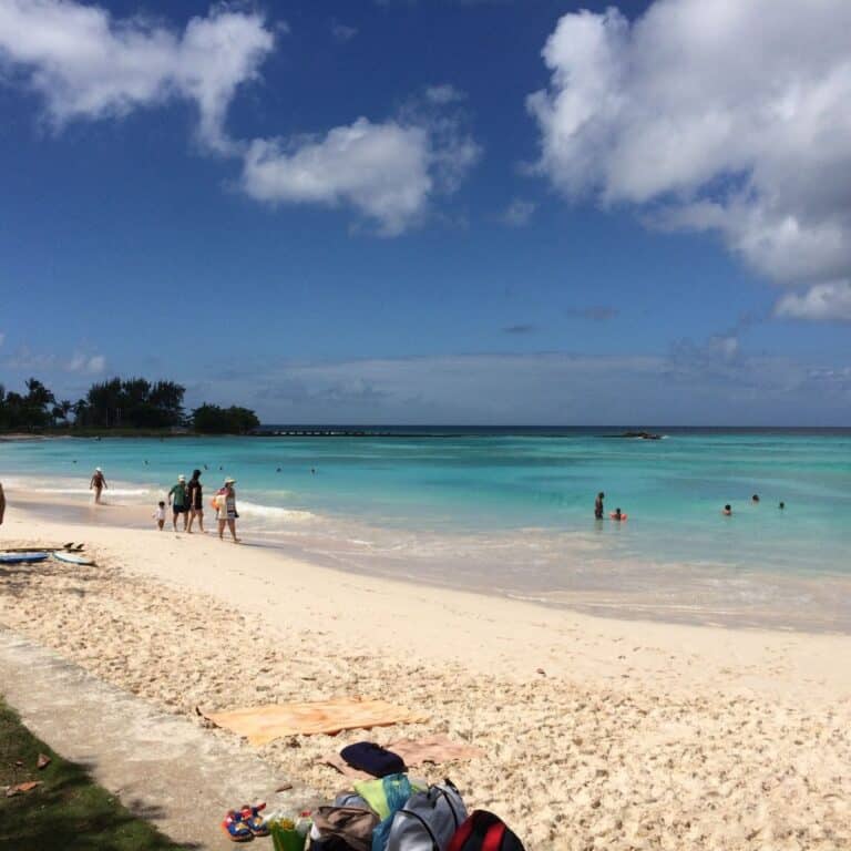 People enjoy a sunny day at a beach with turquoise waters, light sand, and a partly cloudy sky. Towels and bags are on the sand near the shore.