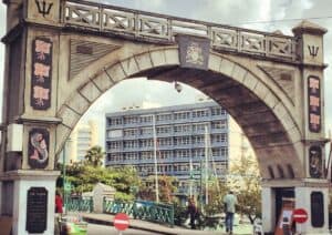 An architectural street arch with decorative panels and a coat of arms stands prominently over a bridge, much like the captivating sights you'll find among things to do in Barbados. Nearby, a modern building and trees are visible under a partly cloudy sky.
