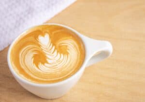 A cup of coffee with intricate latte art sits on a wooden surface next to a white cloth, evoking the serene vibe you'd find if you were wondering, "Is there a Starbucks in Saint Lucia?.