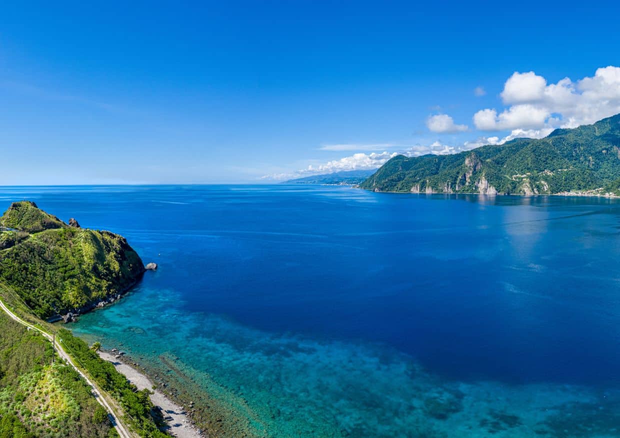 Aerial view of a clear blue ocean meeting rugged green cliffs and lush coastline under a bright blue sky with scattered clouds, reminiscent of the picturesque islands close to St. Lucia.