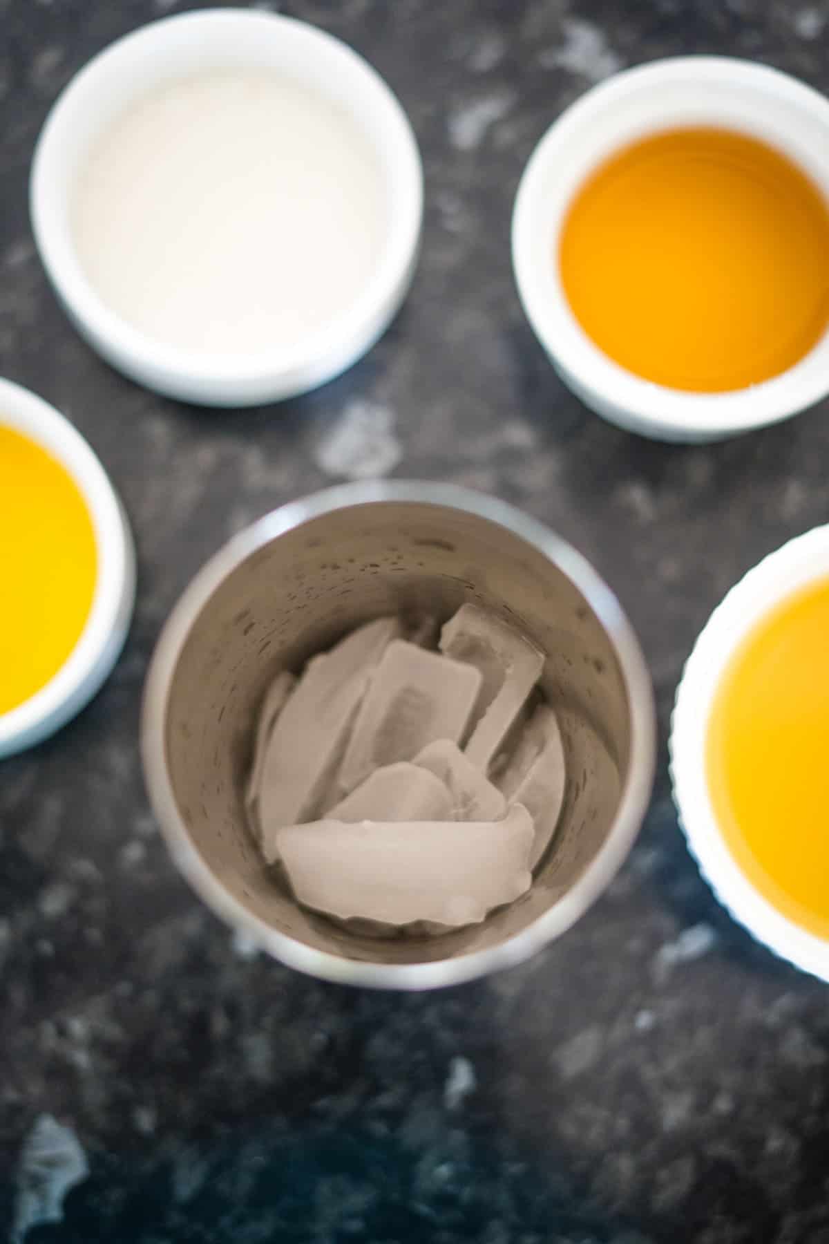 A cocktail shaker filled with ice is surrounded by four small bowls containing various amber and clear liquids on a dark countertop.