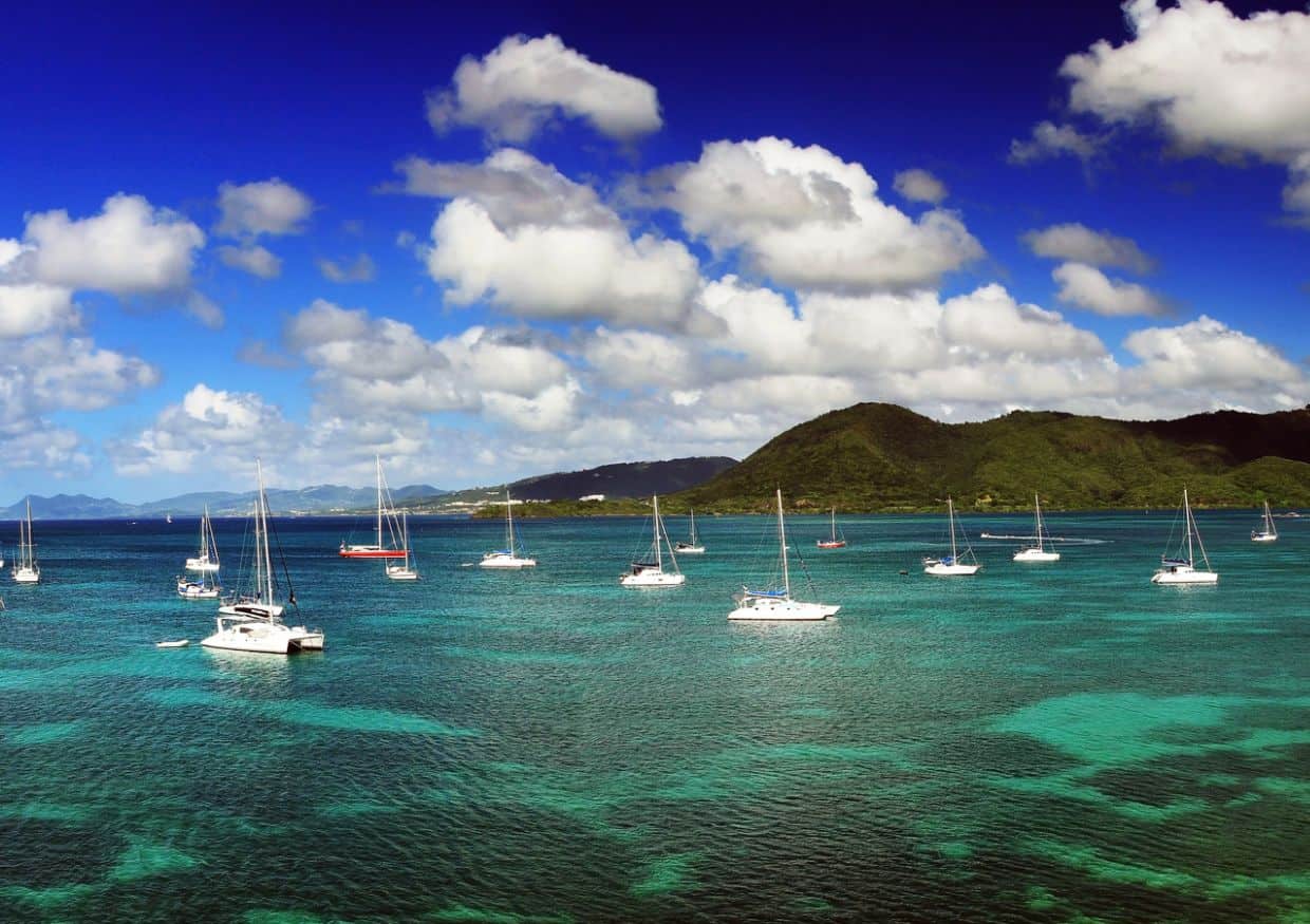 Sailboats drift on turquoise waters beneath a cloudy blue sky, with mountainous islands close to St. Lucia in the background.