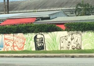 Discover vibrant murals featuring an ice cream truck, a bottle, and a luggage bag with stamps in Barbados. Set against lush foliage and a corrugated building, it's one of the captivating things to do in Barbados.