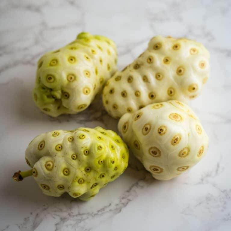 Four noni fruits with bumpy, mottled skin sit on a marble surface.