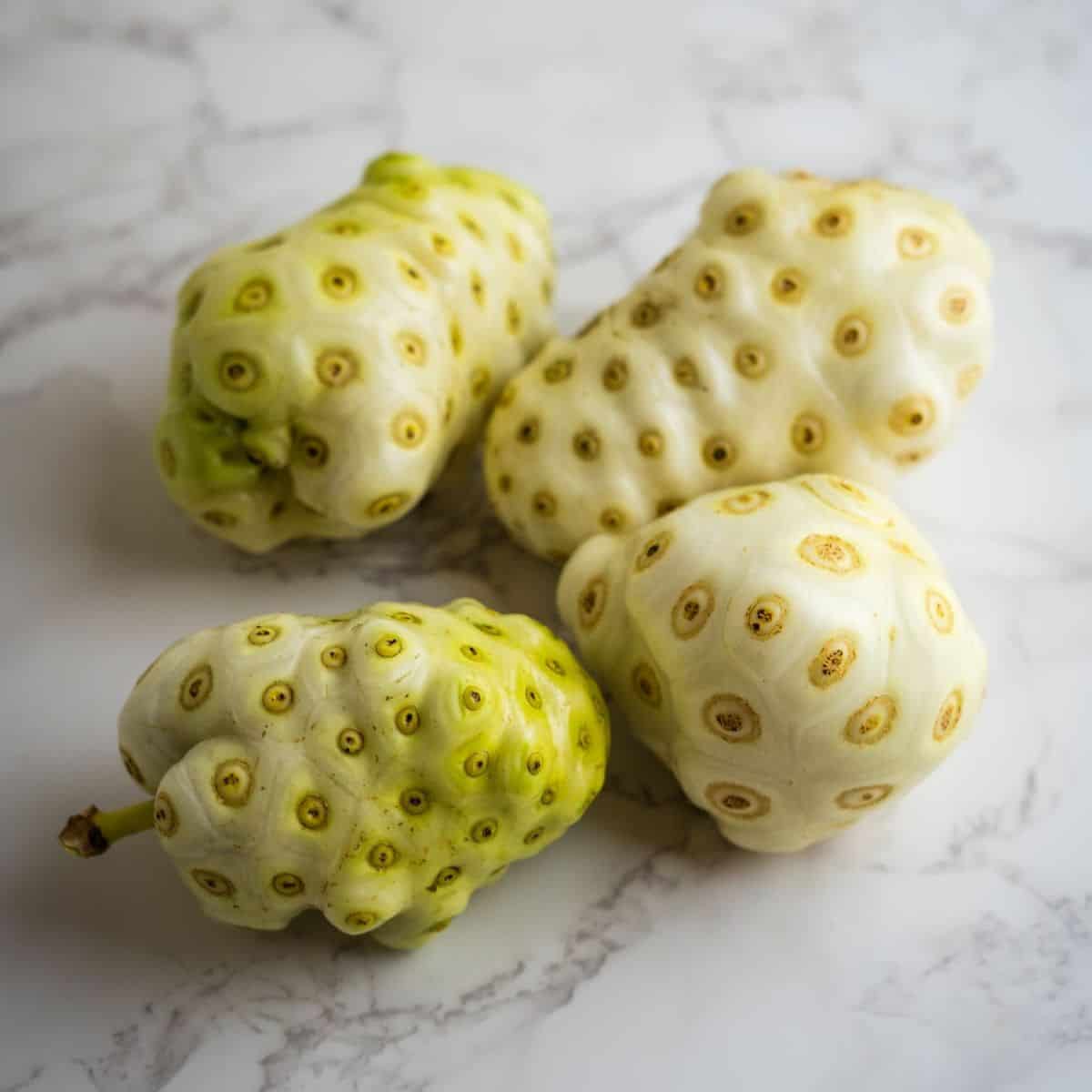 Four noni fruits with bumpy, mottled skin sit on a marble surface.