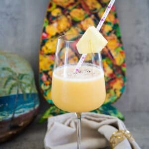 A glass of tropical smoothie garnished with a pineapple slice and straw, in front of a pineapple-shaped art piece.