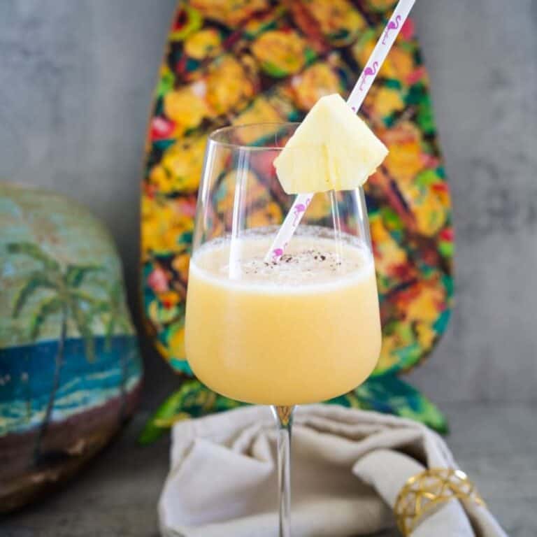 A glass of tropical smoothie garnished with a pineapple slice and straw, in front of a pineapple-shaped art piece.