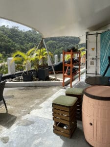 Outdoor patio with a view of trees, offering a hint of adventure akin to a St. Lucia chocolate tour, featuring a canopy, barrel tables, cushioned crate seats, and a red metal shelving unit.