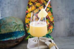 A tropical drink with a pineapple slice on a straw, in a wine glass. A painted coconut and colorful background are in the scene.