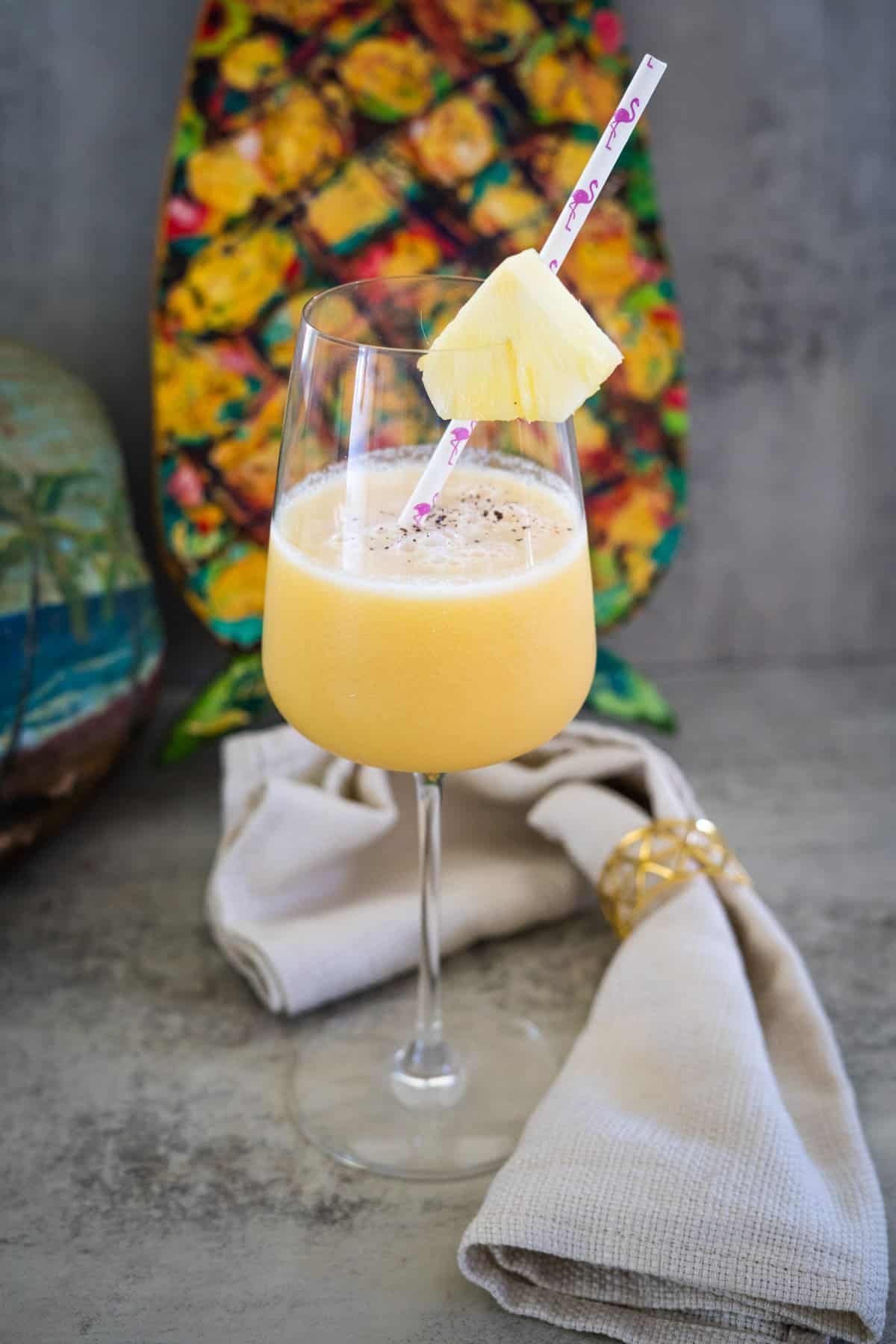 A glass of frothy yellow smoothie with a pink straw and pineapple slice. A colorful background and folded napkin are visible.