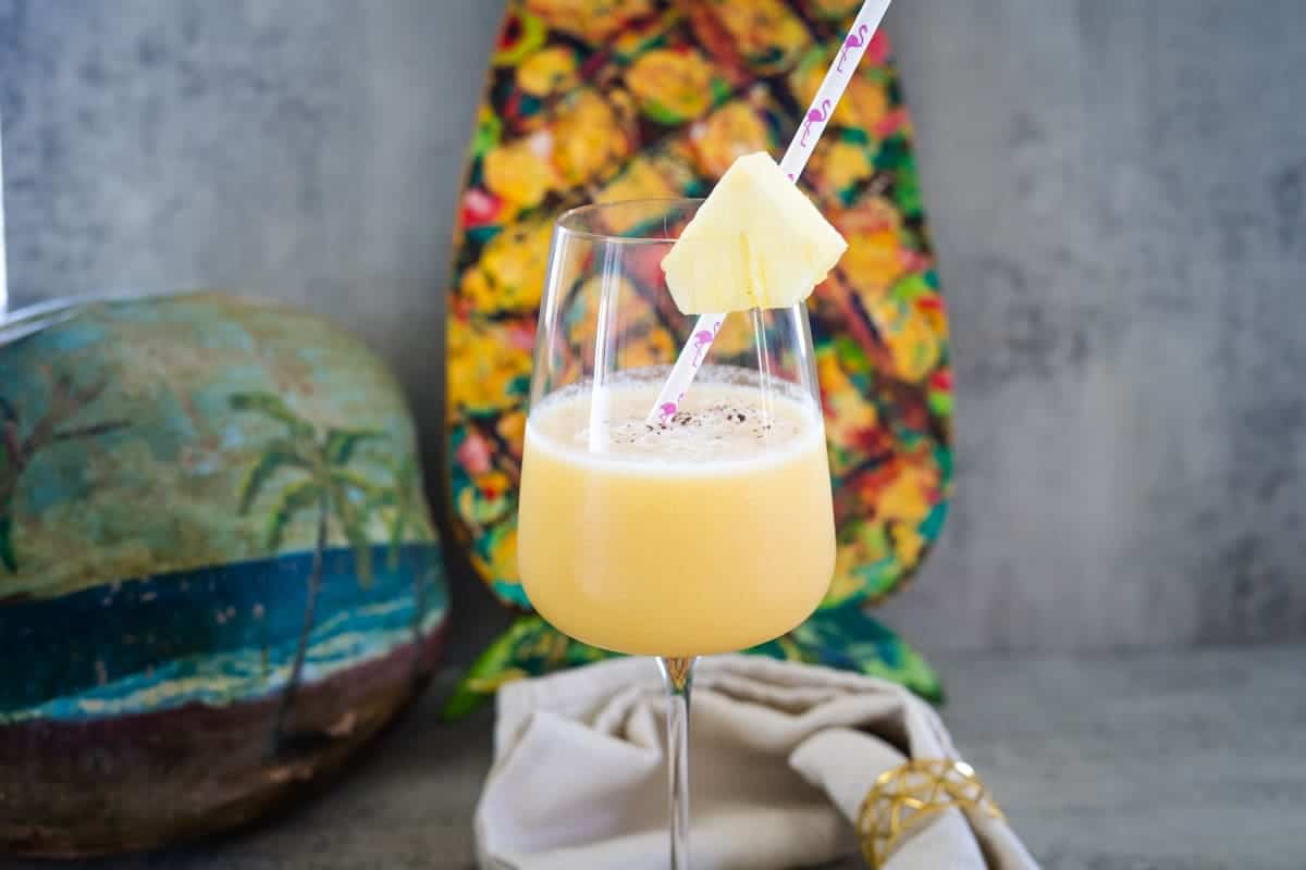 A tropical drink with a pineapple slice on a straw, in a wine glass. A painted coconut and colorful background are in the scene.
