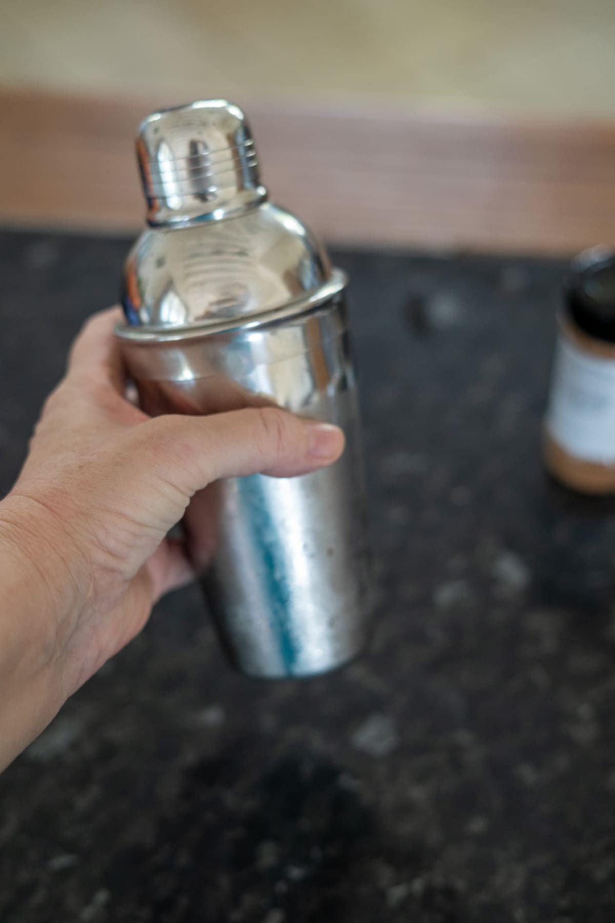 A hand holds a shiny metal cocktail shaker over a dark countertop.