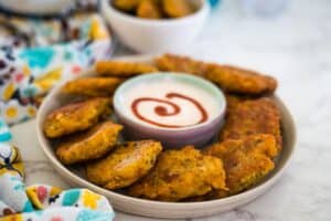 A plate of crisp fried patties encircles a dip with a sauce spiral, evoking curiosity akin to "what is saltfish?". A vibrant cloth adds flair, while a bowl brimming with more golden patties sits invitingly in the background.
