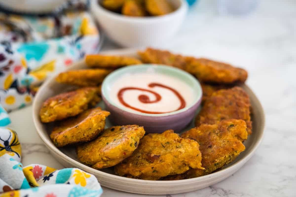 A plate of crisp fried patties encircles a dip with a sauce spiral, evoking curiosity akin to "what is saltfish?". A vibrant cloth adds flair, while a bowl brimming with more golden patties sits invitingly in the background.