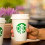 A hand holds a Starbucks coffee cup with the iconic green logo, hinting at the comforting question—"Is there a Starbucks in Saint Lucia?" In the background, a potted plant blooms with vibrant purple flowers.
