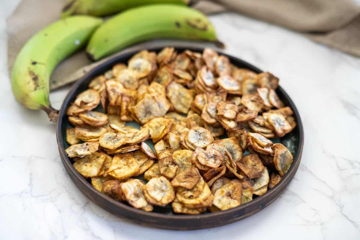 A plate filled with crispy plantain chips next to three unpeeled green plantains on a marble surface.