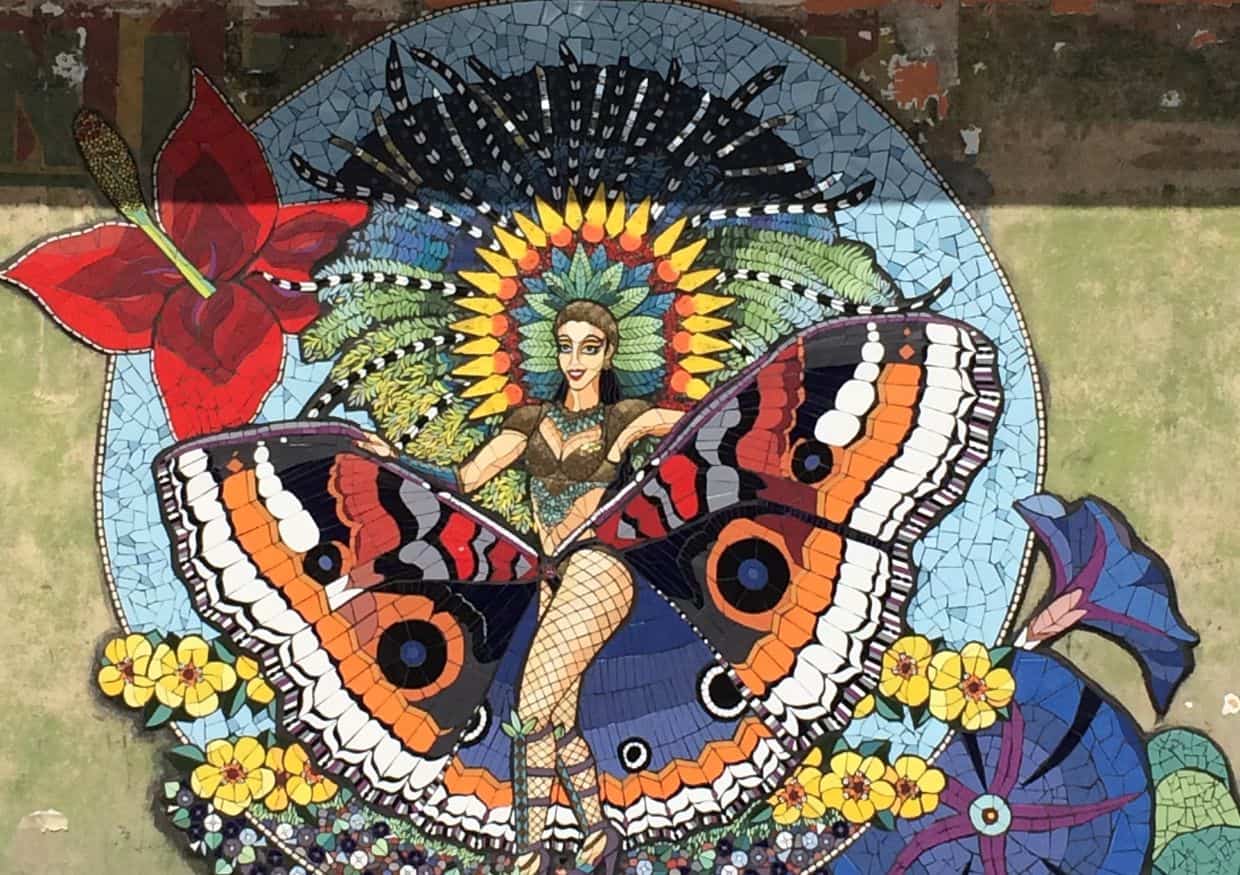 Mosaic mural of a woman with butterfly wings, surrounded by vibrant flowers and colorful patterns in a circular design.