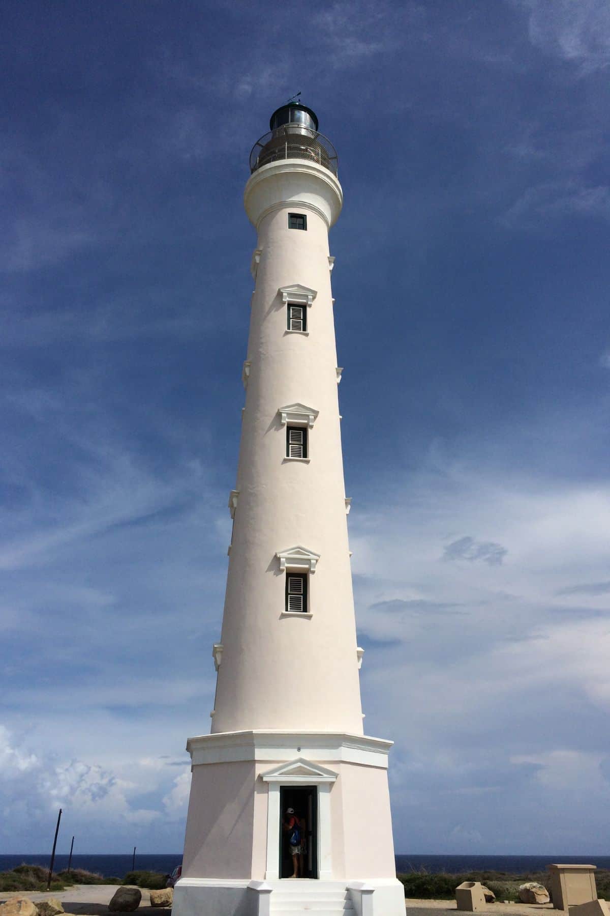 White lighthouse with multiple windows against a blue sky. A person is visible at the base entrance.