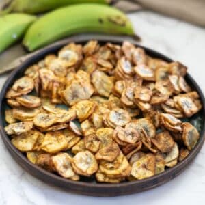 A plate filled with crispy banana chips on a marble countertop, with whole green bananas in the background.