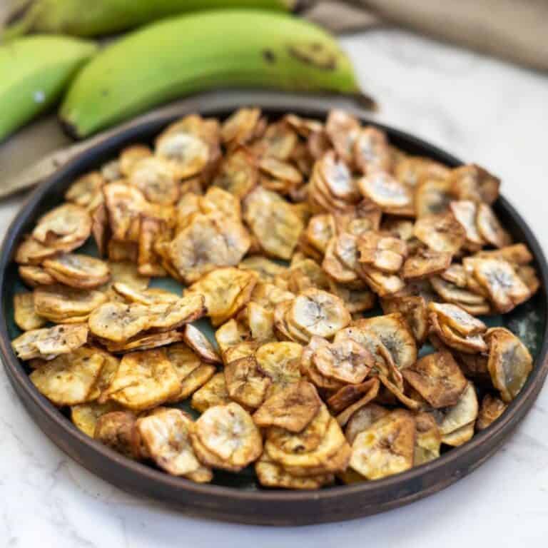 A plate filled with crispy banana chips on a marble countertop, with whole green bananas in the background.