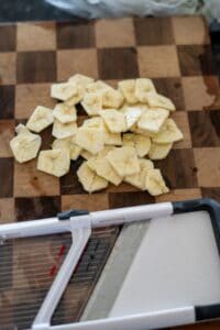 Sliced bananas on a wooden cutting board next to a mandoline slicer, ready to transform into crispy green banana chips.