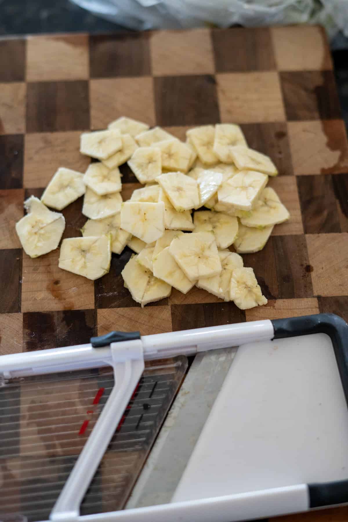 Sliced bananas on a wooden cutting board next to a mandoline slicer, ready to transform into crispy green banana chips.