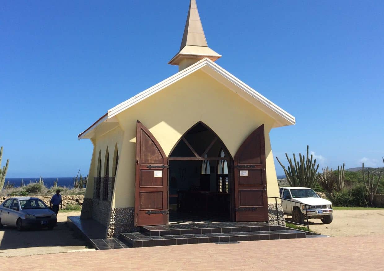 A small, yellow chapel with a steeple and open wooden doors stands surrounded by cacti and parked cars under a clear blue sky. The ocean is visible in the background.