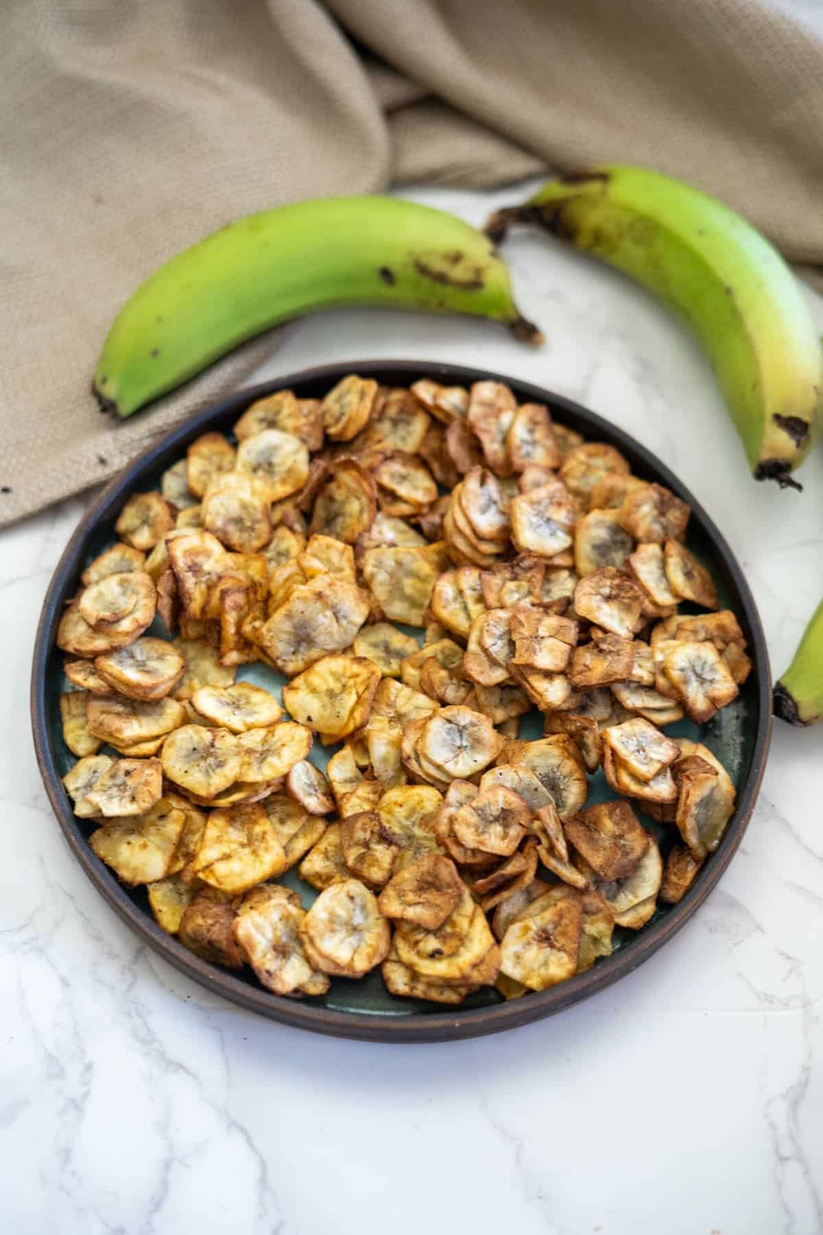 A plate of fried plantain chips is surrounded by three whole unripe green bananas on a marble surface. A tan cloth is partially visible in the background.