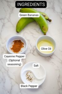 On a marble surface, green bananas, destined to become crispy banana chips, are displayed alongside olive oil, optional cayenne pepper, salt, and black pepper. Each ingredient is thoughtfully labeled.