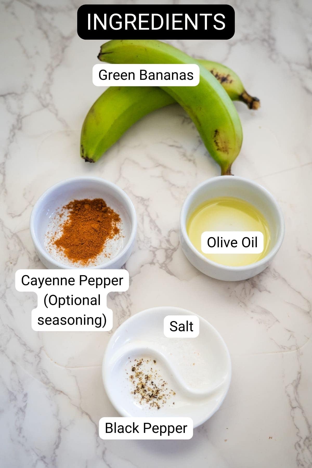 On a marble surface, green bananas, destined to become crispy banana chips, are displayed alongside olive oil, optional cayenne pepper, salt, and black pepper. Each ingredient is thoughtfully labeled.