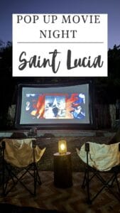 Outdoor pop-up cinema setup in Saint Lucia with a large screen playing a film. Two chairs and a lantern add charm to the scene. A sign above reads "Pop Up Movie Night.