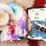 Open suitcase with denim shorts, a tropical-print shirt, vintage camera, pink sunglasses, a sun hat, and a palm tree-patterned garment on a carpeted floor.