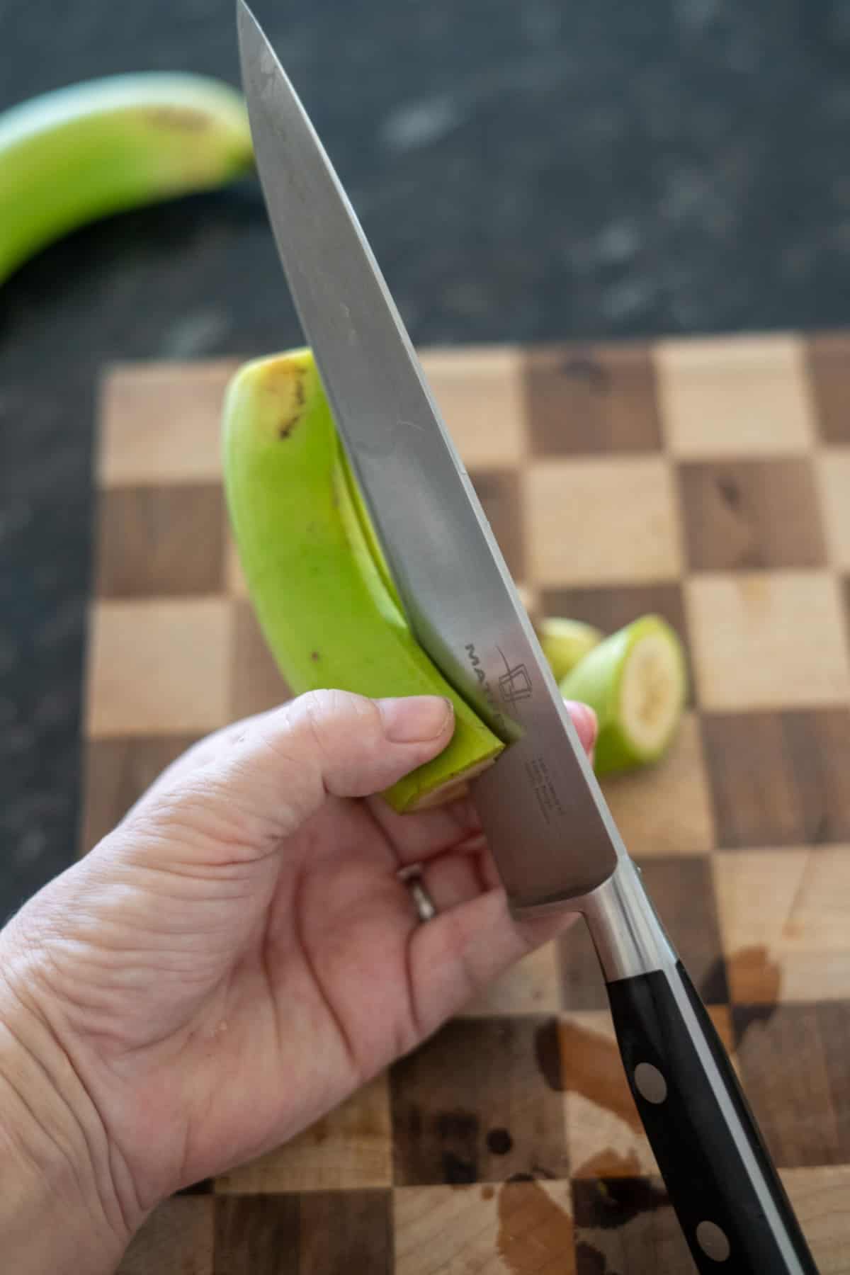 A hand precisely slices an unpeeled plantain on a wooden cutting board, preparing the first step for making crisp green banana chips.