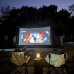 Outdoor movie setup with two empty chairs facing an inflatable screen showing a film. A small lantern on a wooden table is placed between the chairs. Trees create a silhouette in the background.