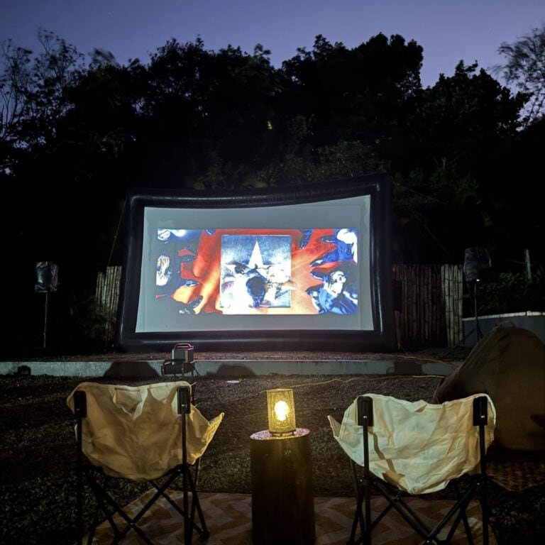 Outdoor movie setup with two empty chairs facing an inflatable screen showing a film. A small lantern on a wooden table is placed between the chairs. Trees create a silhouette in the background.