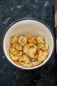 A bowl filled with green banana chips and sliced plantains seasoned with spices sits enticingly on a dark countertop.
