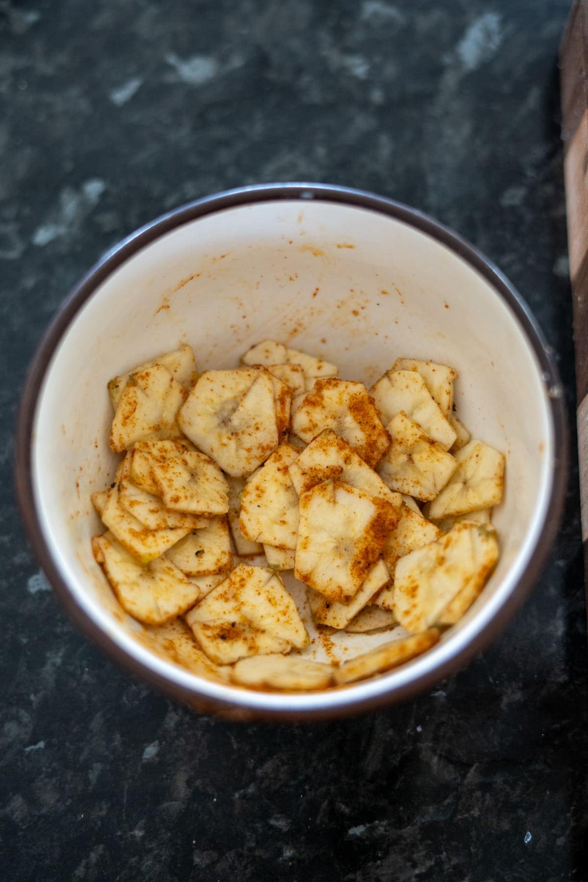 A bowl filled with green banana chips and sliced plantains seasoned with spices sits enticingly on a dark countertop.